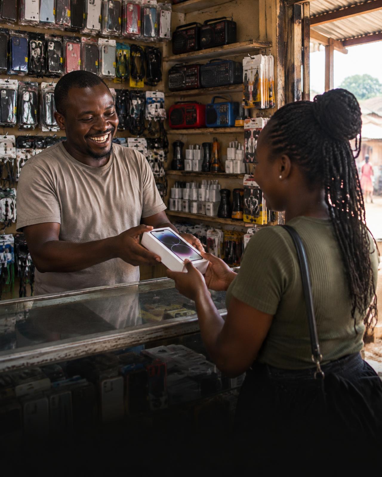A shopkeeper hands a boxed phone to a smiling customer in a Kampala electronics shop